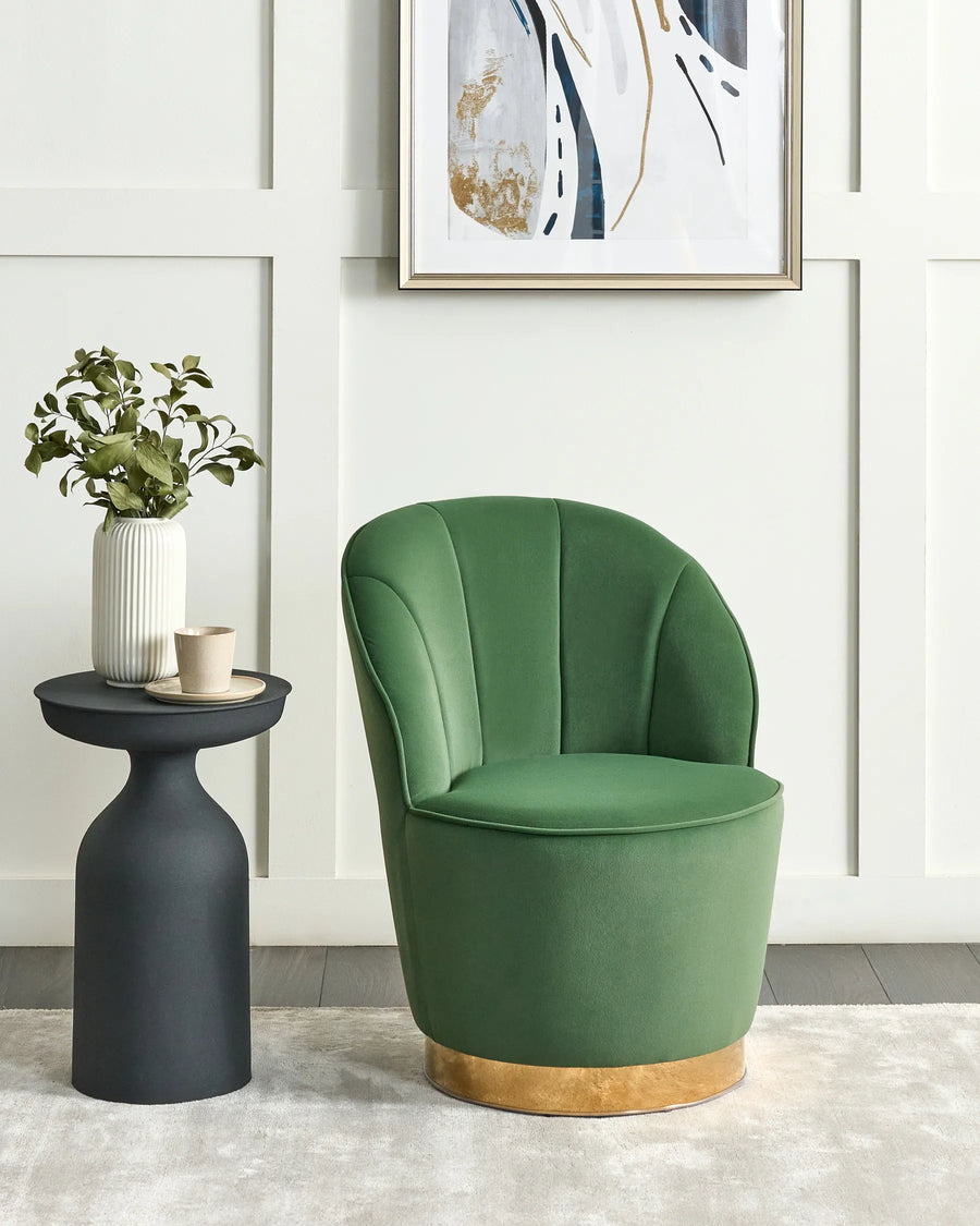 Green armchair with gold base next to a black side table with a plant, in a room with white walls and a framed abstract painting.