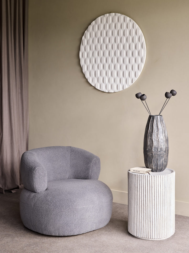 Gray armchair in a room with a textured wall mirror and a white side table with a vase.