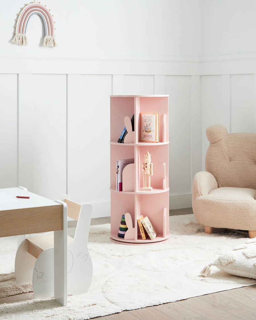 Pink children's bookshelf with books in a room with a white wall, beige chair, and wooden floor.