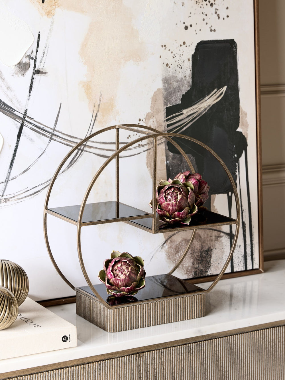 Decorative table setting with a gold circular shelf and artichoke sculptures against an abstract wall.