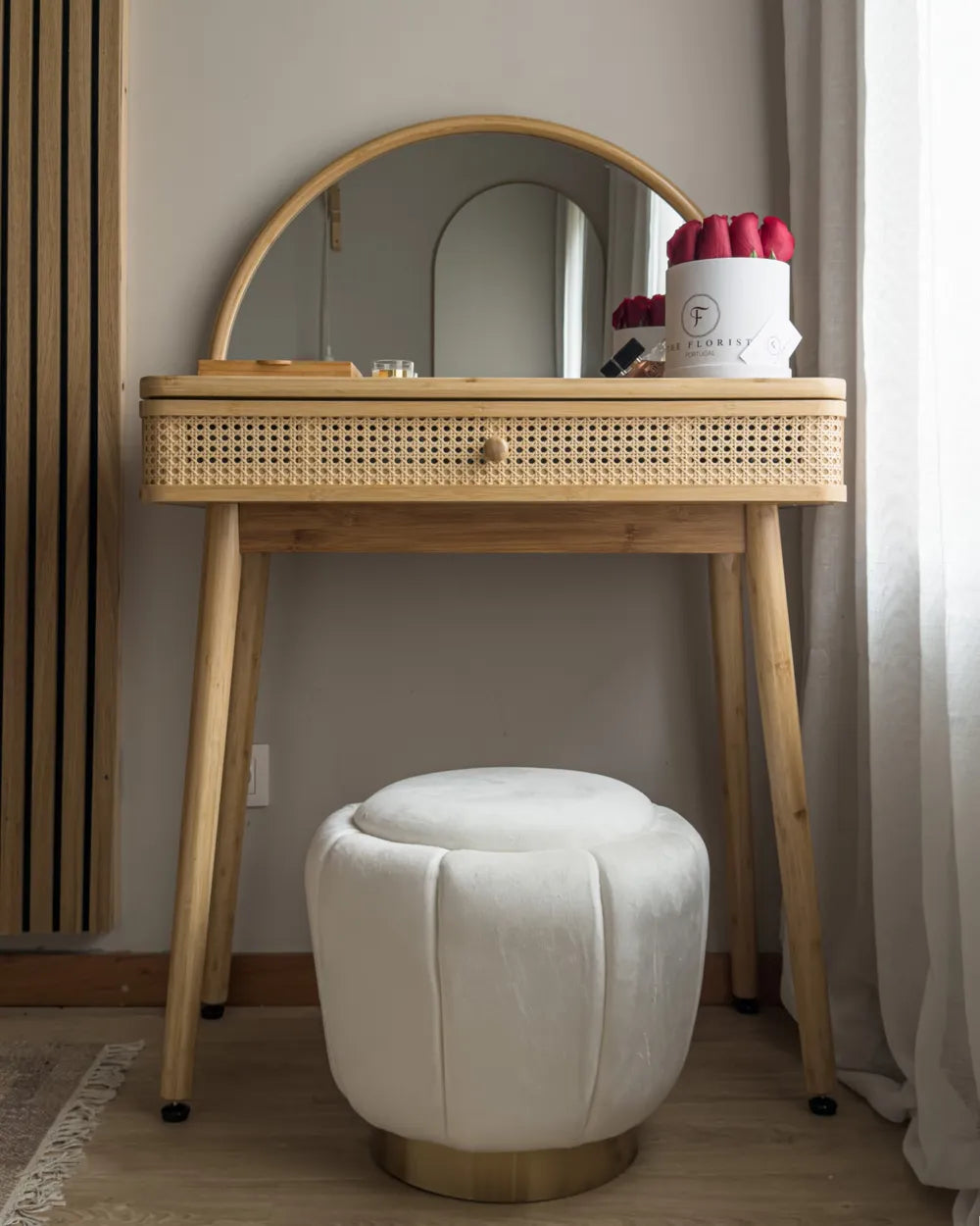 A light natural wood vanity table with a woven cane drawer front and an arched mirror, paired with a round white velvet stool in a softly lit room.