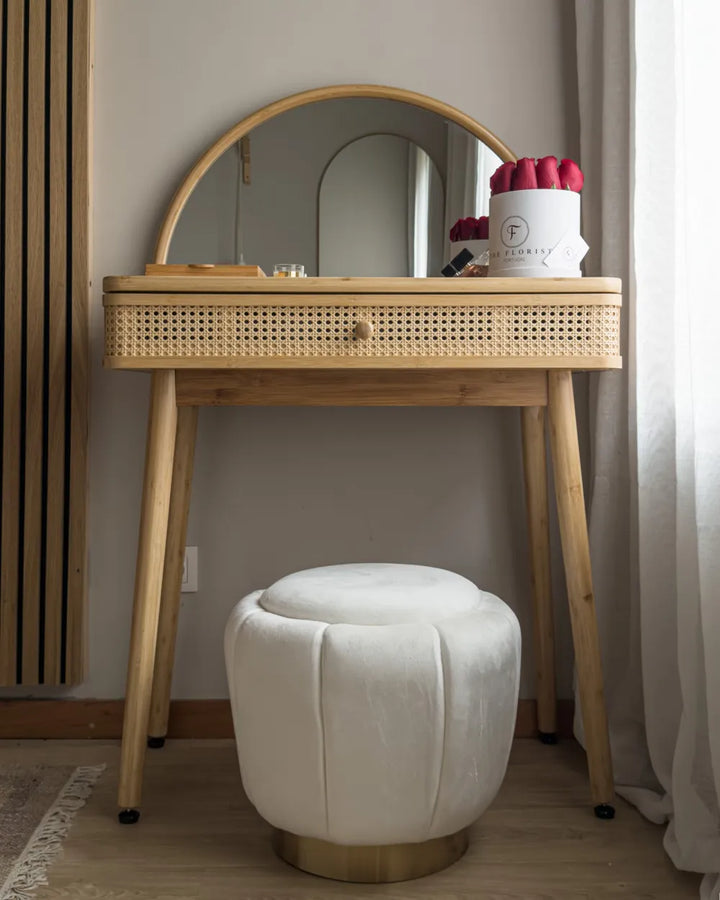 A light natural wood vanity table with a woven cane drawer front and an arched mirror, paired with a round white velvet stool in a softly lit room.