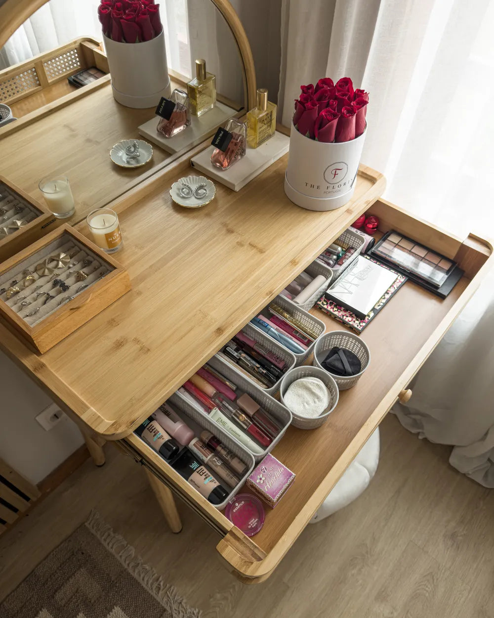 A light natural wood makeup vanity table with an open lower drawer filled with cosmetics, shown in a bright room with curtains and a mirror above.