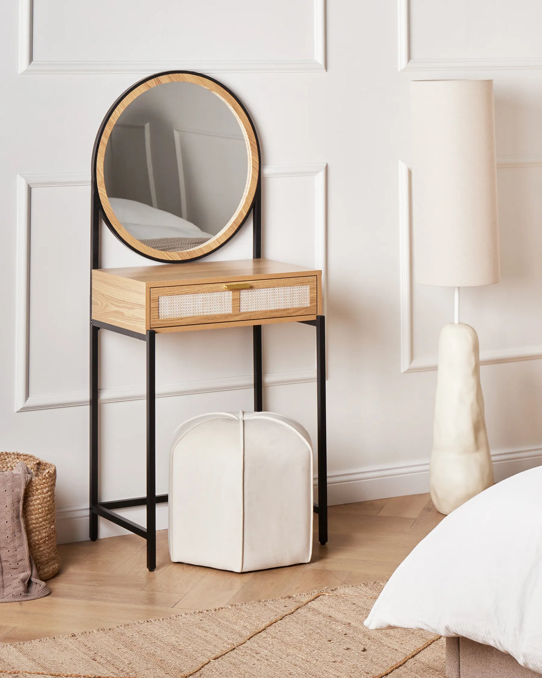 Small wooden side table with round mirror next to a white ottoman and lamp in a bedroom setting.