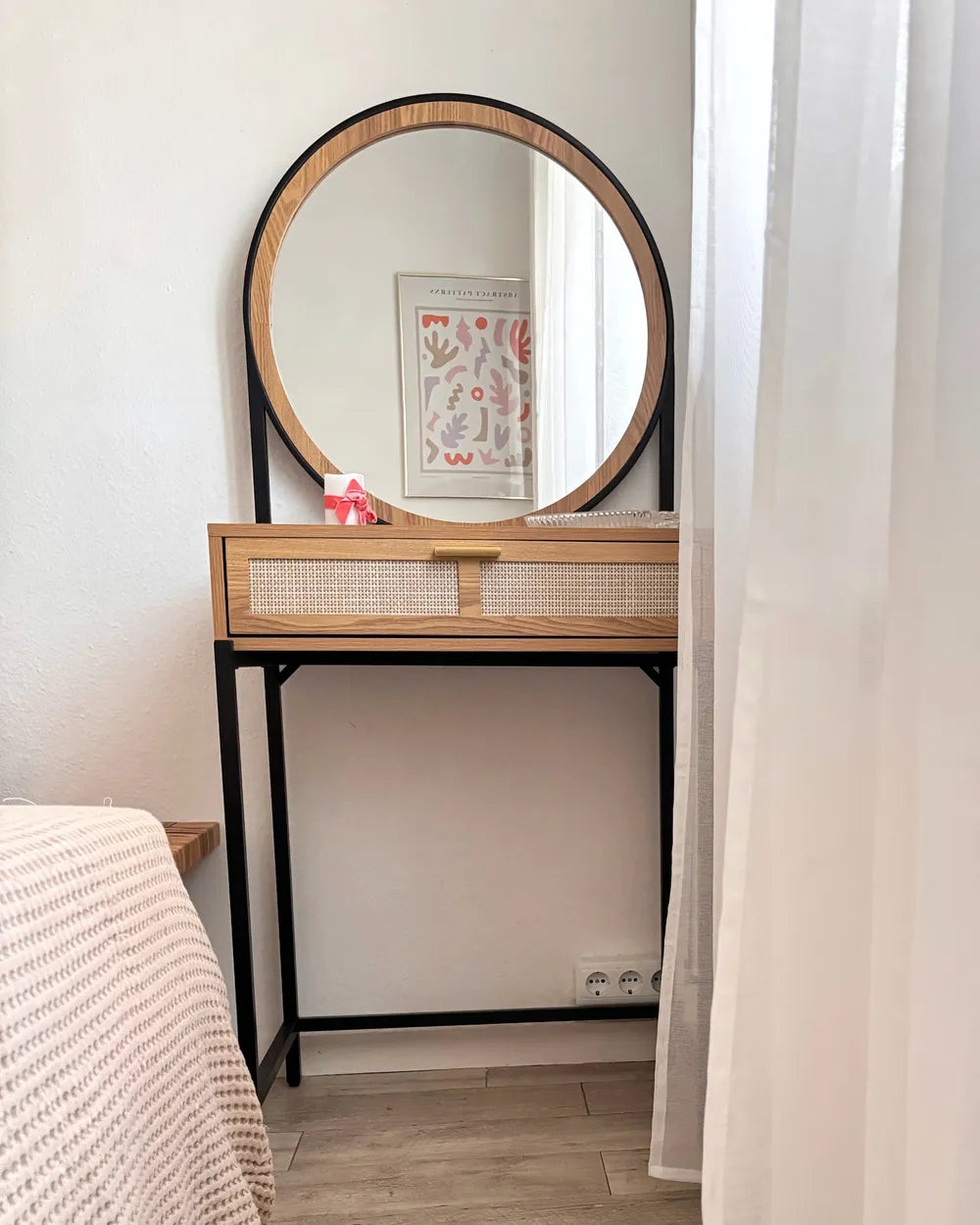 A modern vanity table with a large round mirror, featuring a light wood frame, woven cane drawer front, and black metal legs, set in a bedroom corner.