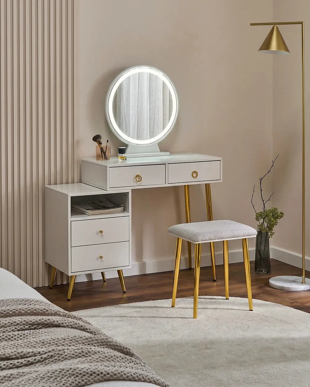 A modern vanity table set in a bedroom, featuring a white vanity with gold legs, an illuminated round mirror, a matching stool, and a side cabinet.