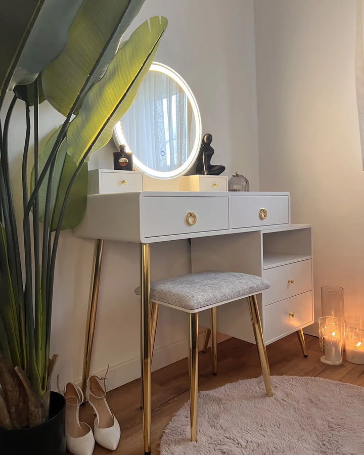 A white vanity table with gold legs and gold drawer knobs, featuring a round backlit mirror, a matching upholstered stool, and styled in a warm room with a potted plant, candles, and shoes.
