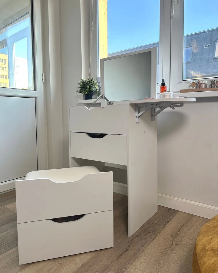 A white vanity desk with an attached mirror and fold-out shelf, paired with a matching two-drawer stool, shown in a bright room with wood flooring and large windows.