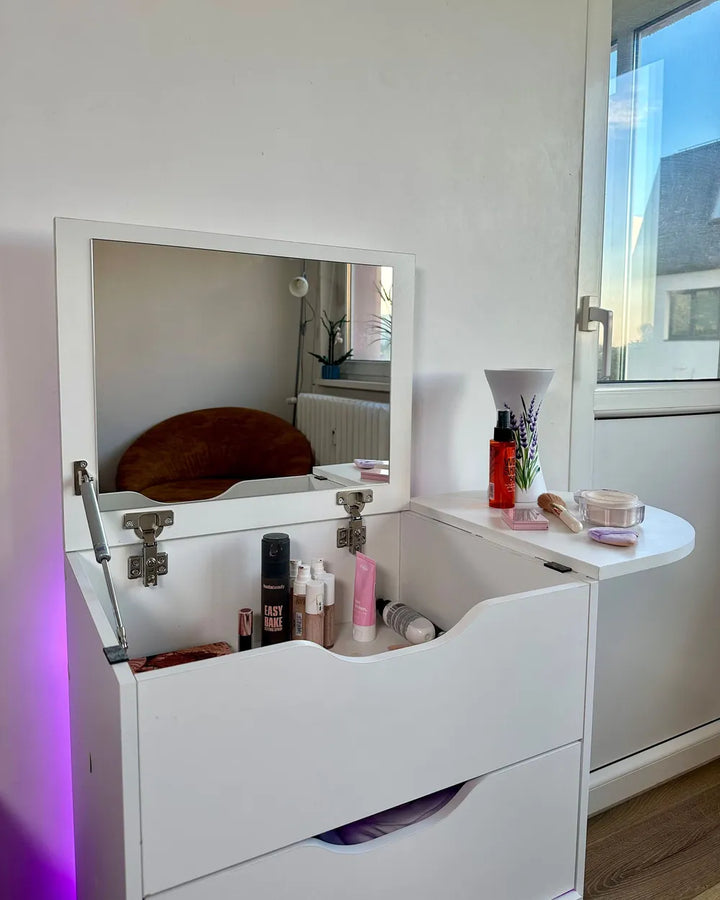 A white vanity desk with an open mirror and a hinged side table, styled with various cosmetic bottles and jars in a room setting.