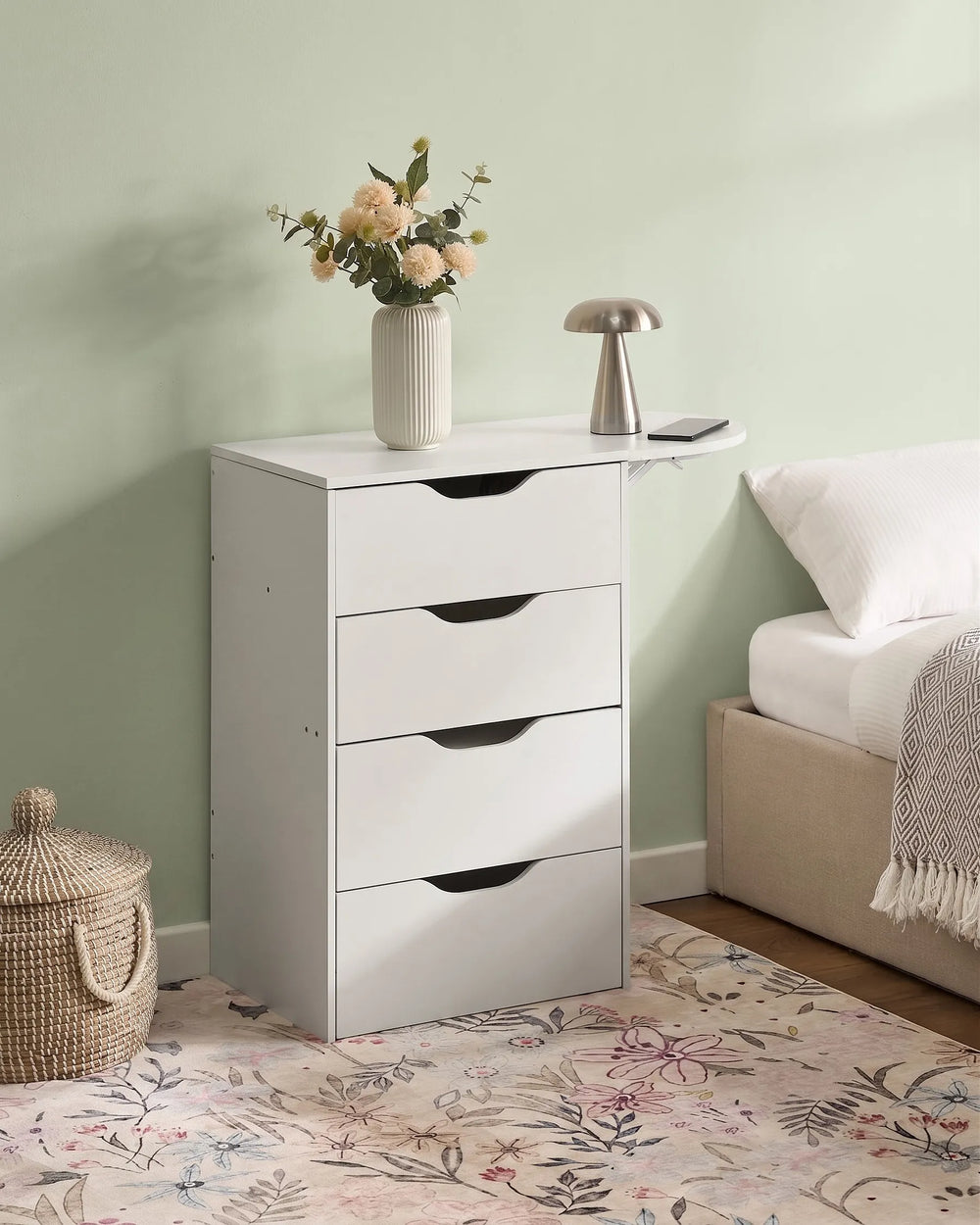 A white four-drawer nightstand with a small attached side table and shelf, styled in a bedroom next to a bed with a vase of flowers and a metallic lamp on top.