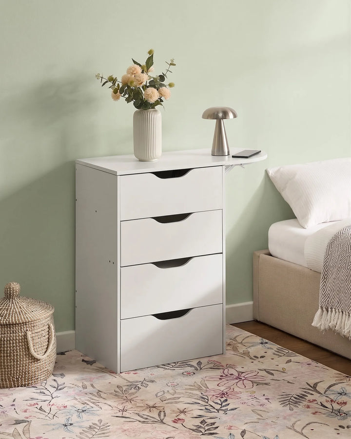 A white four-drawer nightstand with a small attached side table and shelf, styled in a bedroom next to a bed with a vase of flowers and a metallic lamp on top.