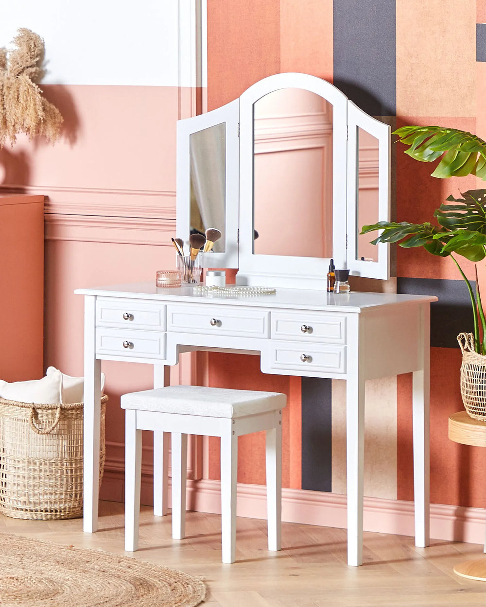 White vanity table with three-panel mirror and matching stool in a styled room with pink and peach walls, a plant, and woven decor.