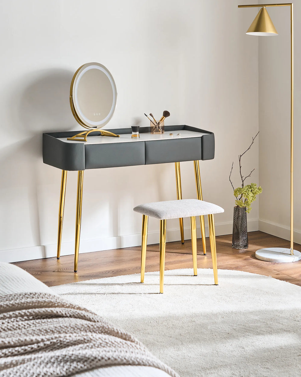 A modern vanity table with a gray upholstered top, gold metal legs, and an attached oval mirror, styled in a bedroom setting with a matching stool, floor lamp, and wood flooring.