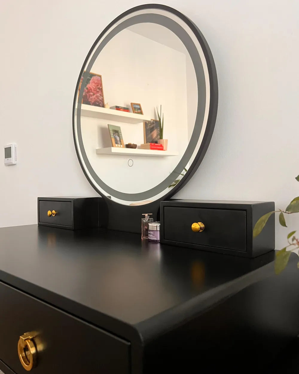 A black vanity table with a large oval mirror, two small matching side drawers, and gold-tone hardware, shown against a white wall.