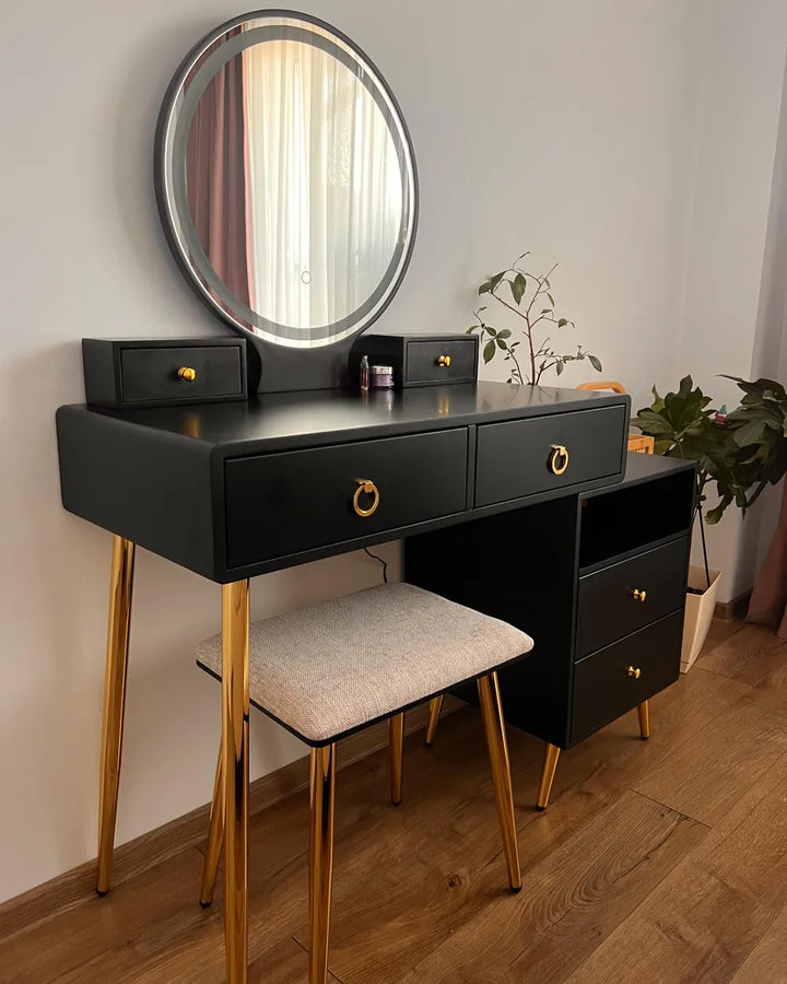 A black vanity table with gold metal legs and drawer pulls, paired with a matching stool and a large round mirror, set in a room with a wood floor and light gray wall.