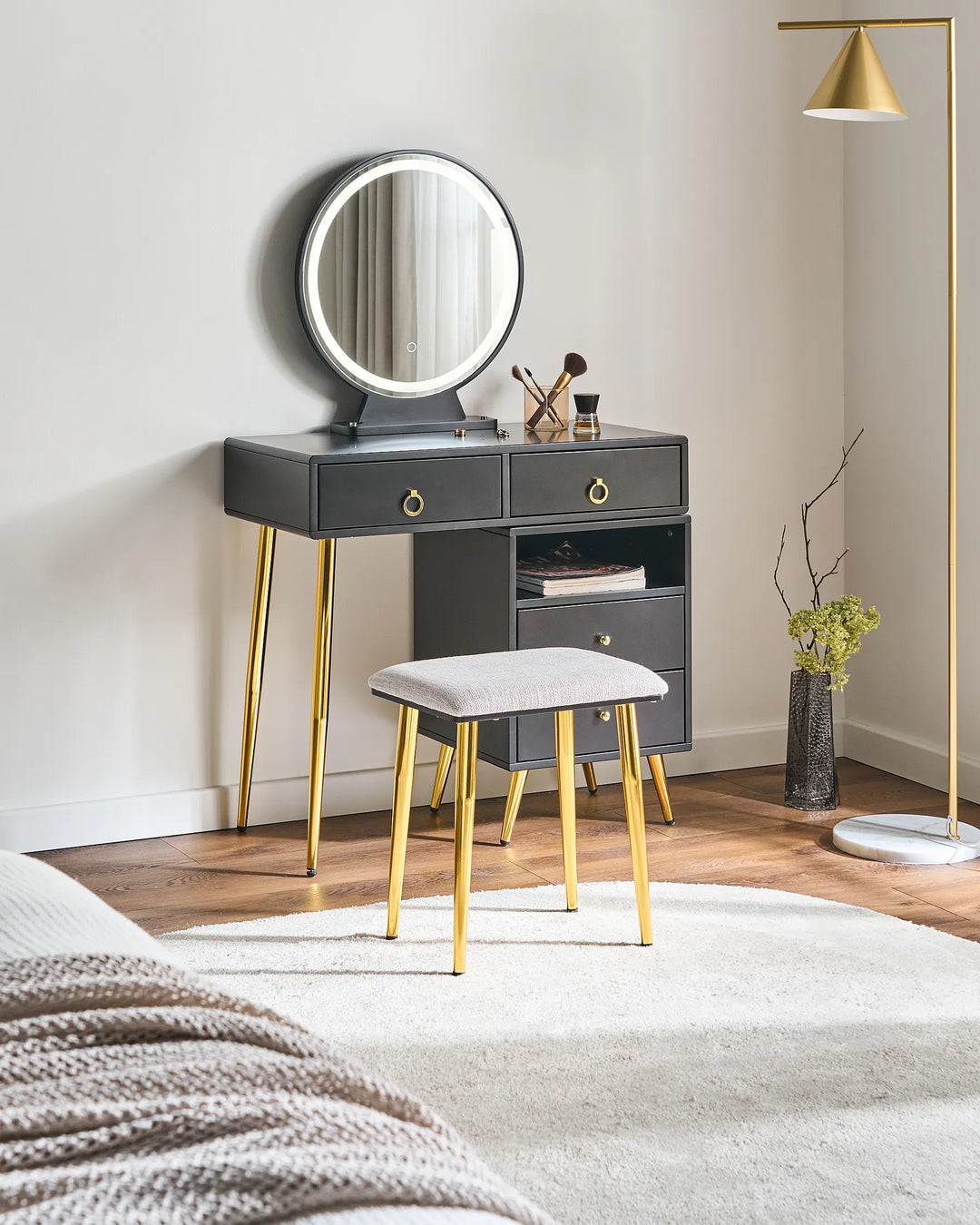 A modern vanity table set in a bedroom, featuring a dark gray console table with gold legs, a round backlit mirror, a matching gray stool with gold legs, and a gold floor lamp nearby.