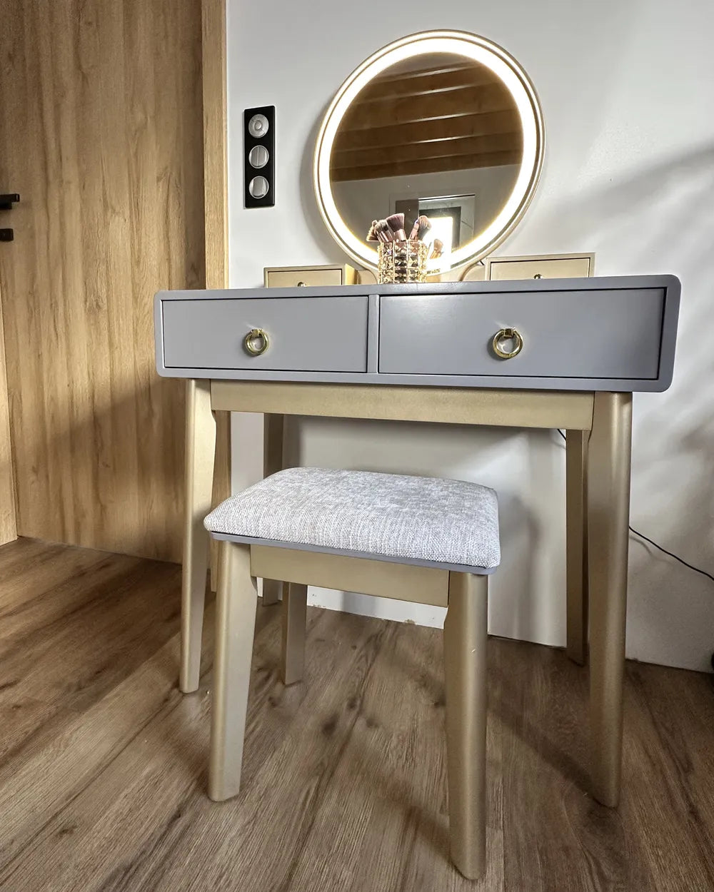 A light gray and natural wood vanity table set with a matching cushioned stool and an illuminated round mirror, shown in a home interior setting.