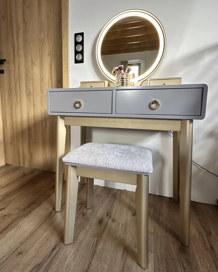 A light gray and natural wood vanity table set with a matching cushioned stool and an illuminated round mirror, shown in a home interior setting.