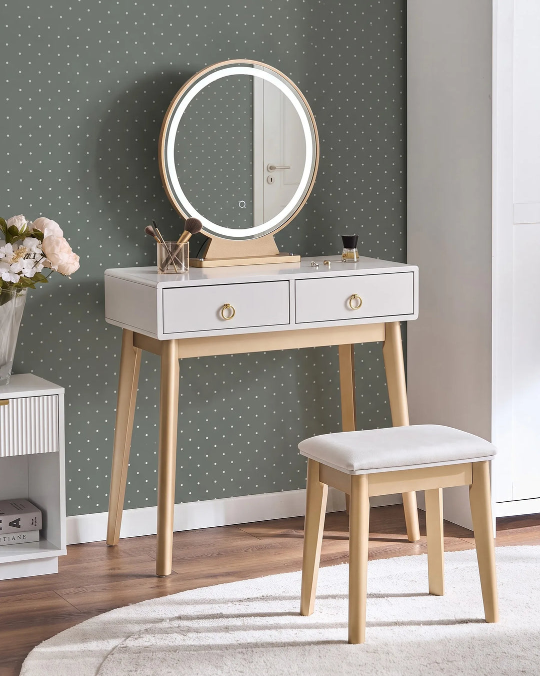 A modern vanity table set with a white drawer-front console, light wood legs, a round backlit mirror, and a matching cushioned stool, styled in a room with green polka-dot wallpaper.