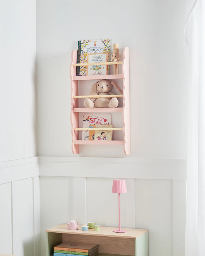 Pink wall shelf with books and a teddy bear in a room with a lamp and books on a table.
