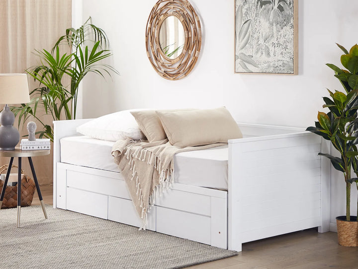 White daybed with beige pillows and blanket in a room with plants and decor.