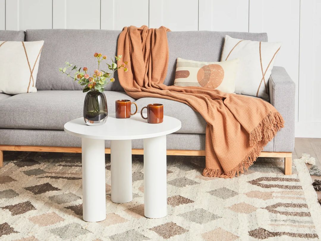 Gray sofa with a brown throw blanket, white side table with mugs and flowers, and patterned rug.