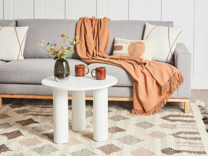 Gray sofa with a brown throw blanket, white side table with mugs and flowers, and patterned rug.