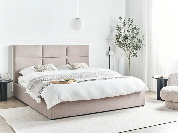 Beige upholstered bed in a modern bedroom setting with white walls and minimal decor.