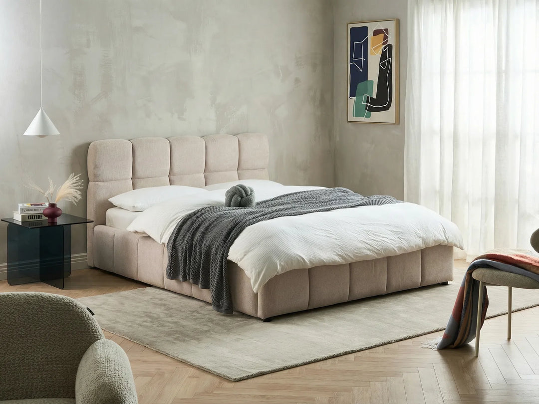 Beige upholstered bed in a modern bedroom with a side table and artwork on the wall.