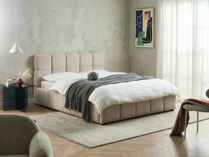 Beige upholstered bed in a modern bedroom with a side table and artwork on the wall.