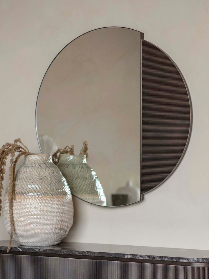Round mirror with a textured black half on a neutral wall, next to a textured white vase.