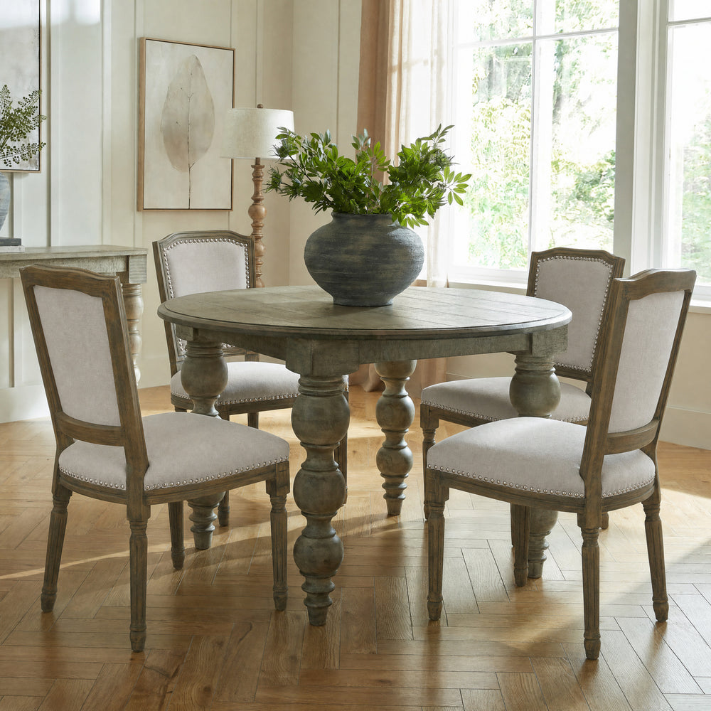 A round distressed gray wooden dining table with four matching upholstered chairs in a bright, sunlit dining room. A large dark blue-gray ceramic vase filled with green foliage sits on the table.