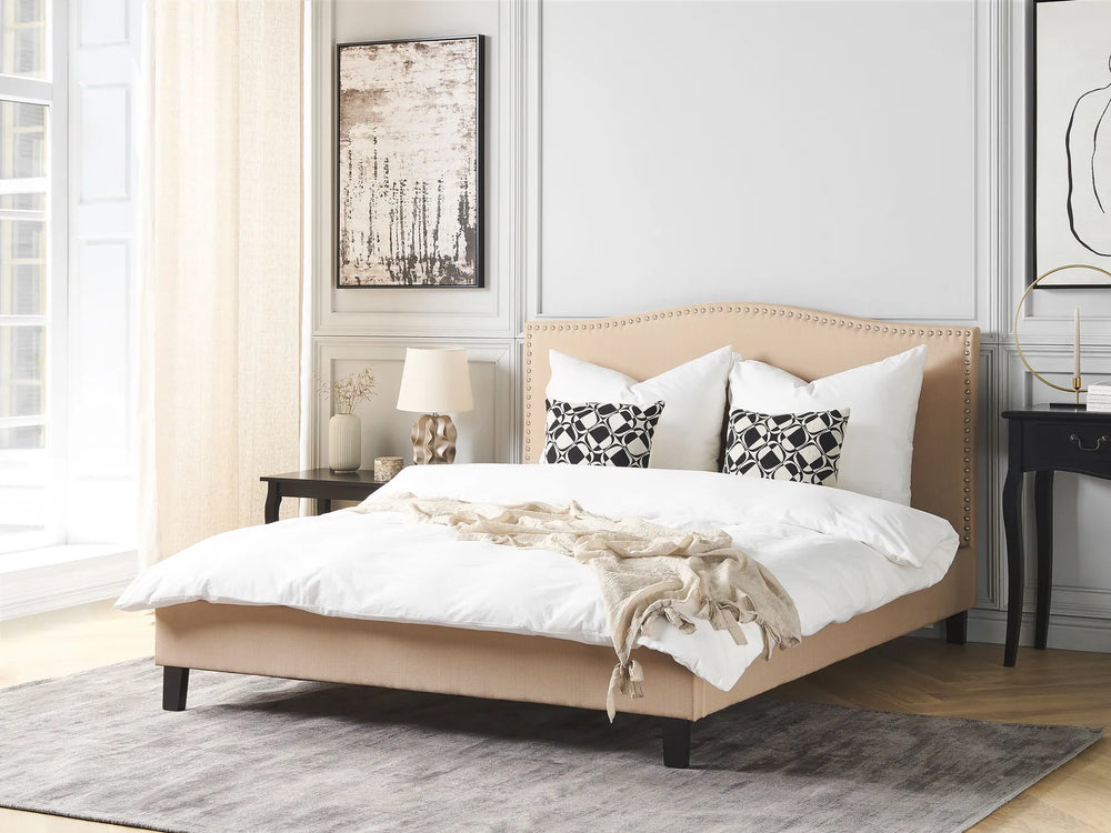 Beige upholstered bed with white bedding and black and white patterned pillows in a bedroom setting.