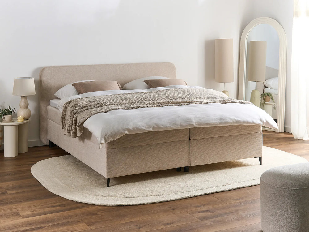 Beige bed in a bedroom setting with wooden flooring and white walls.