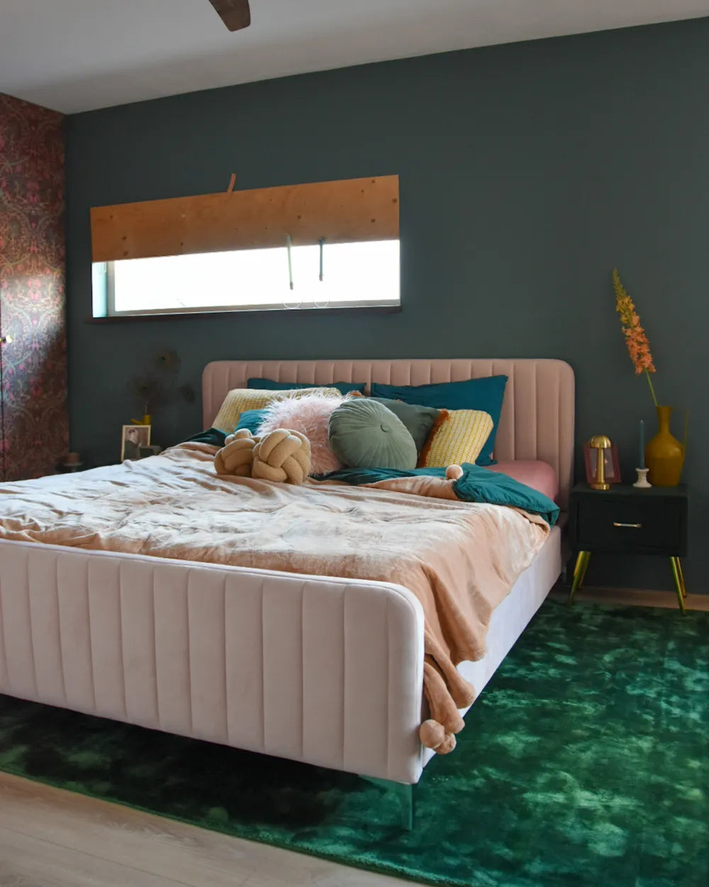 Neatly made bed with colorful pillows in a bedroom with dark walls and a green rug.