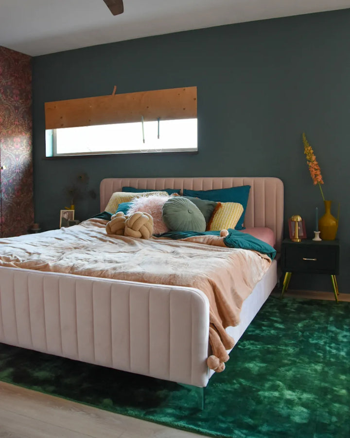 Neatly made bed with colorful pillows in a bedroom with dark walls and a green rug.