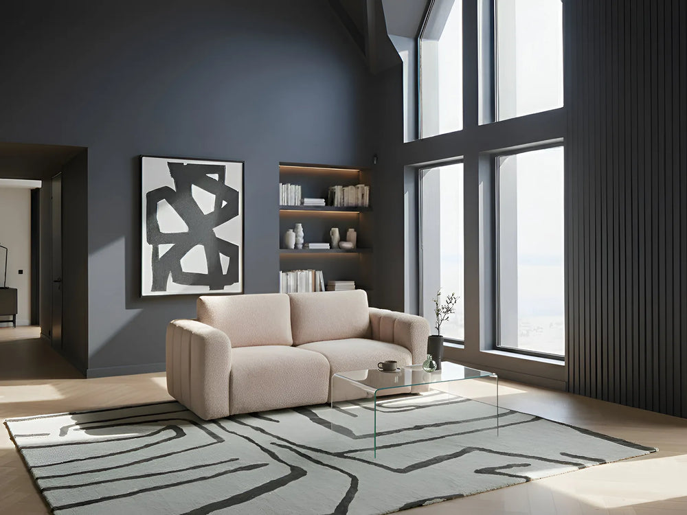 Modern living room with a beige sofa, glass coffee table, and large windows.