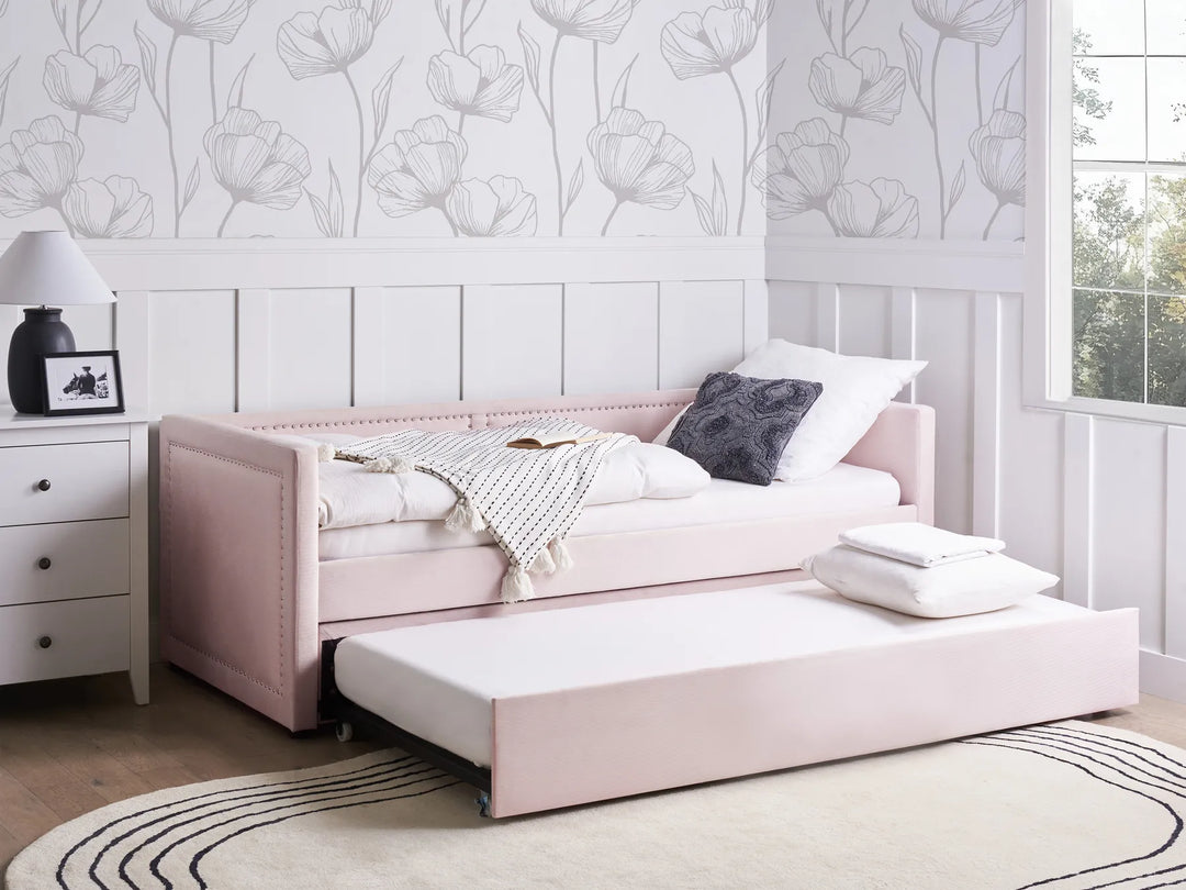 Pink sofa bed with pull-out trundle in a room with floral wallpaper and a lamp.