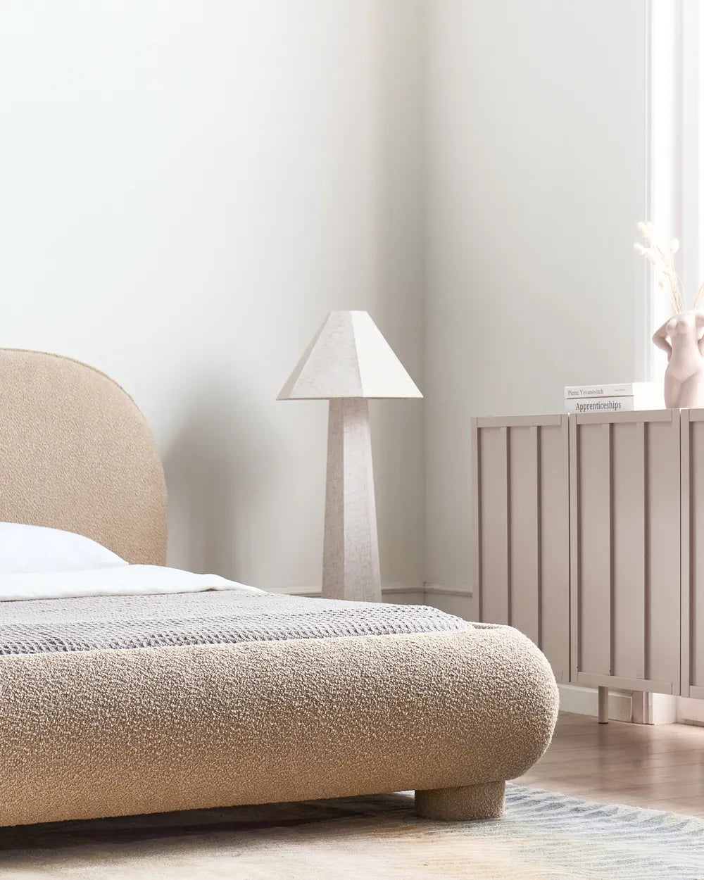 Beige upholstered bed in a minimalistic bedroom with a lamp and radiator.