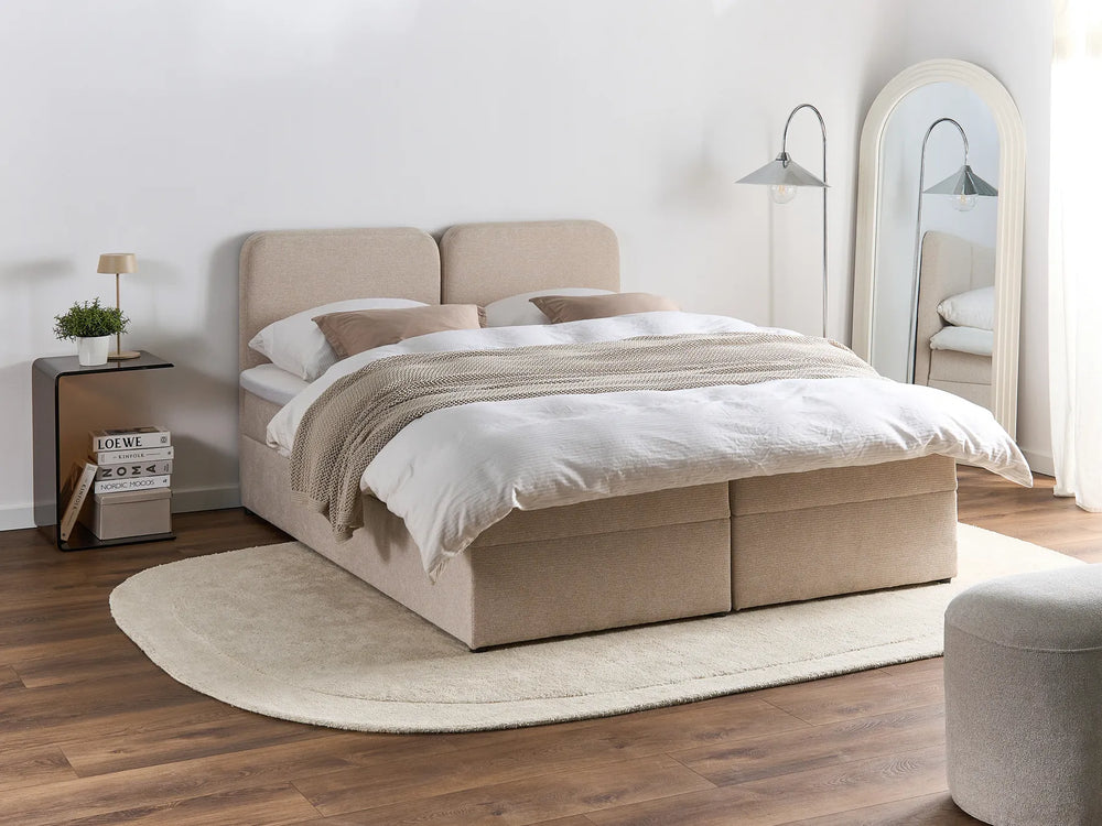 Beige upholstered bed in a bedroom setting with wooden flooring and a mirror.
