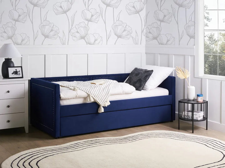 Blue upholstered bed with white bedding in a room with floral wallpaper and a window.
