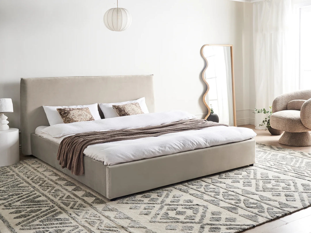 Beige upholstered bed in a modern bedroom with a patterned rug and decorative mirror.