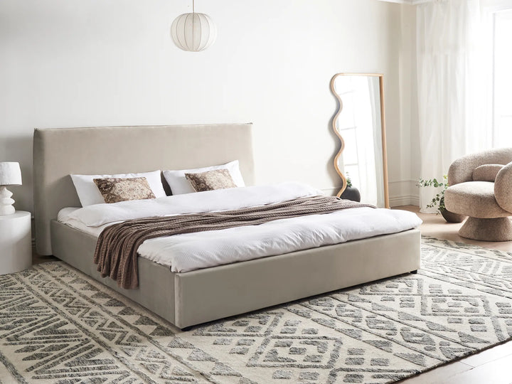 Beige upholstered bed in a modern bedroom with a patterned rug and decorative mirror.