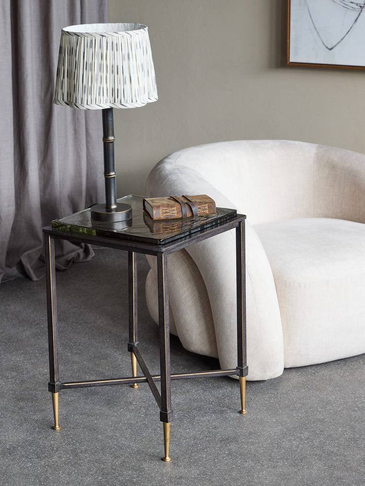 Small side table with a lamp and decorative items next to a white armchair in a room.
