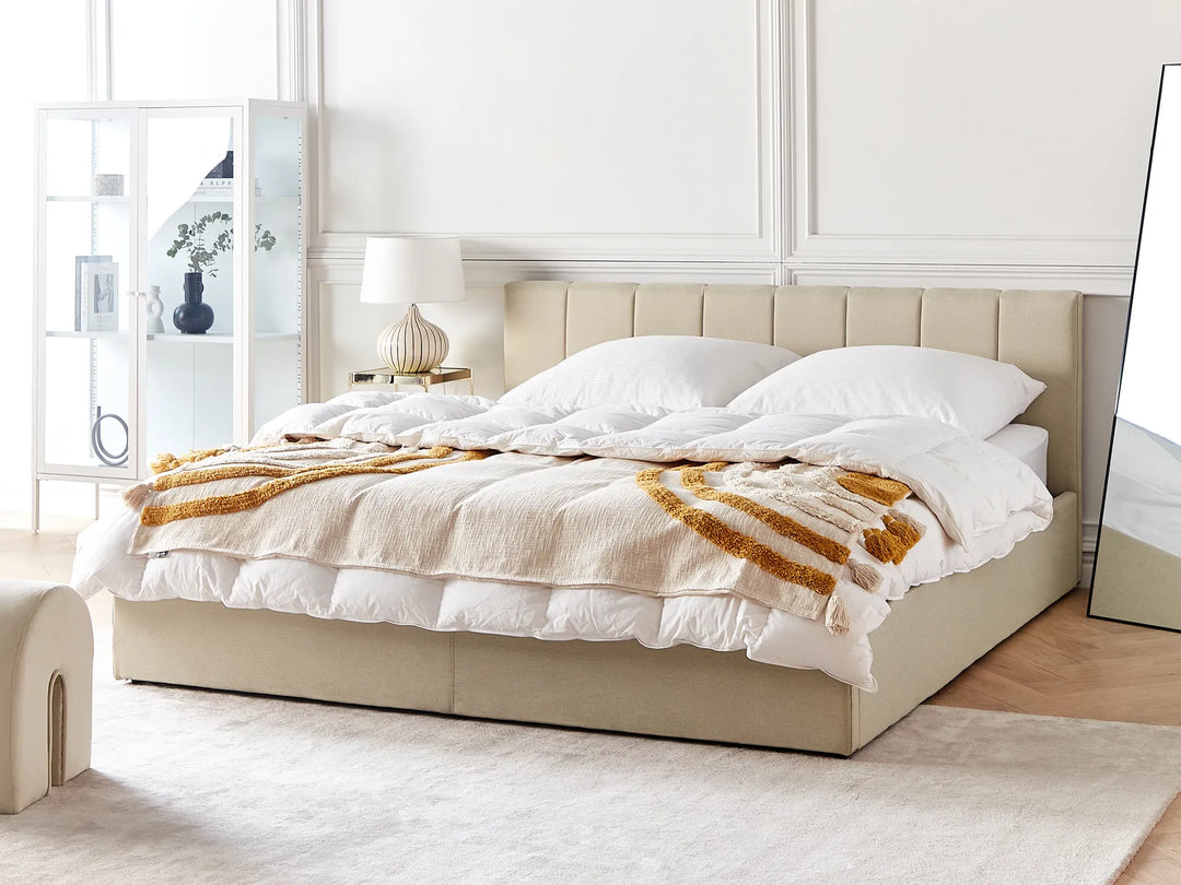 Beige upholstered bed with white bedding and a gold throw blanket in a bright bedroom.