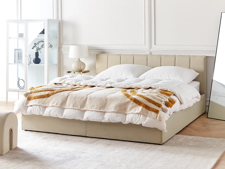 Beige upholstered bed with white bedding and a gold throw blanket in a bright bedroom.