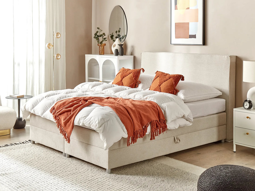 Beige upholstered bed with white bedding and orange throw in a bedroom setting.