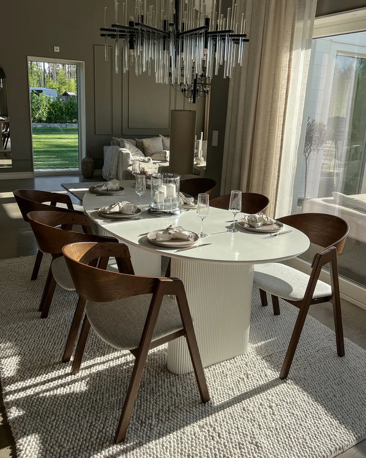 Modern dining room with a round table and wooden chairs, featuring a chandelier and large windows.