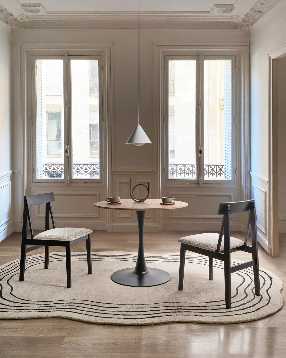 Black dining chairs with cream upholstered seats styled around a small round table in a bright, minimalist dining space with elegant moldings, tall windows, and neutral décor.