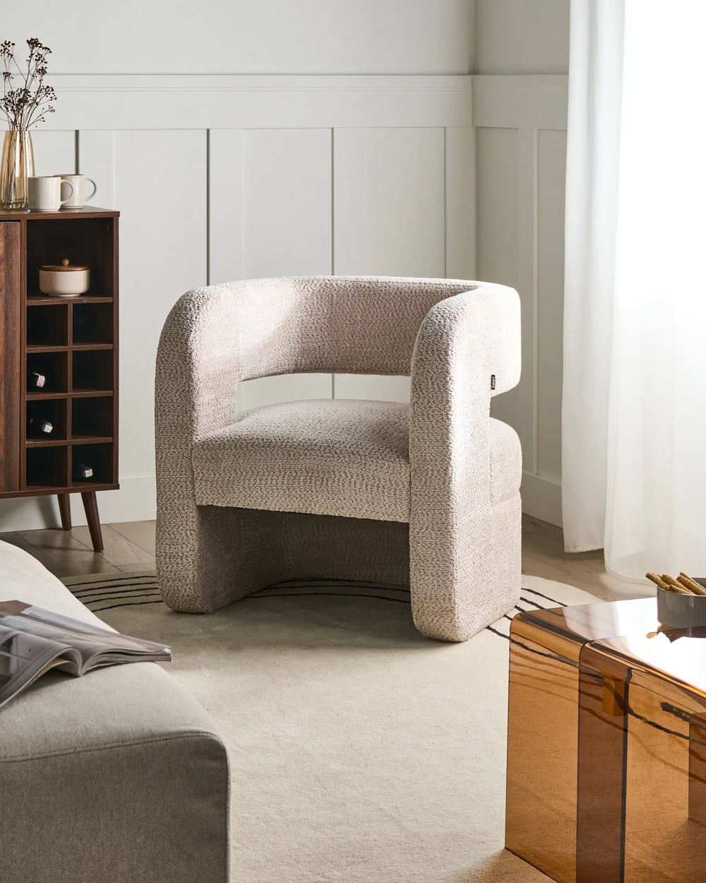 Beige armchair in a modern living room setting with a wooden side table and bookshelf.